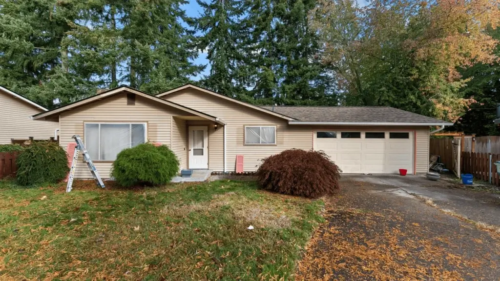 Single-story beige house with attached garage, white front door, two large bushes in front yard, a ladder by the left window, and tall evergreen trees in the background. Driveway has scattered autumn leaves.