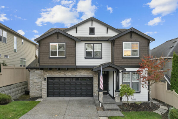 Two-story modern house with brown and white siding, black garage door, stone accent, and a small front porch. An American flag is by the entrance, and there’s a small front yard with landscaping.