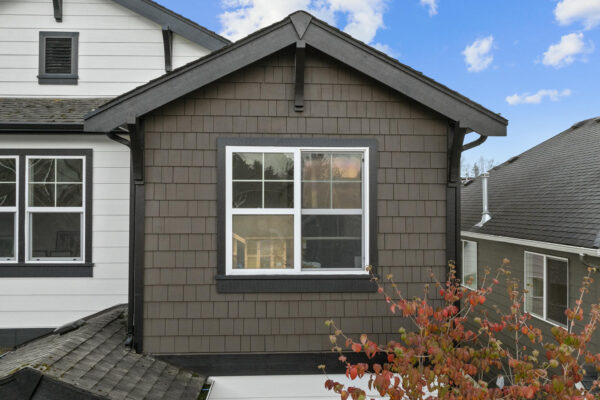 Close-up of the upper floor of a house with brown shingle siding, a white-framed window, and a gray roof. Adjacent houses and a tree with reddish leaves are visible. Blue sky with a few clouds in the background.