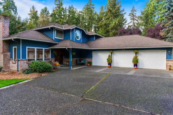 A two-story blue house with white trim, a brick chimney, and a three-car garage. The driveway is large and bordered by plants. The house is surrounded by tall green trees and has potted plants by the entrance.