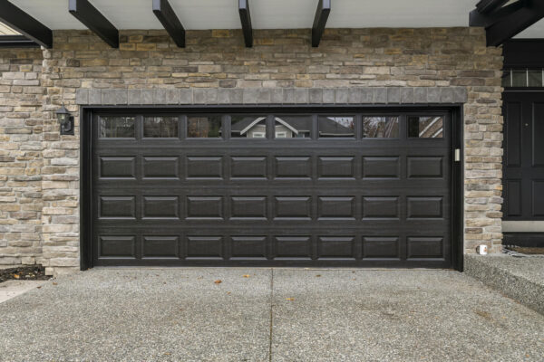 A modern black garage door with rectangular panels and windows at the top, set in a stone exterior wall next to a black front door and a concrete driveway.