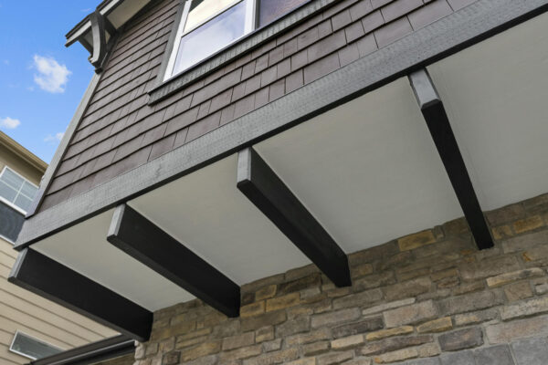 Close-up view from below of a house exterior showing dark exposed beams, stone wall, brown shingle siding, and a window, with blue sky and clouds in the background.