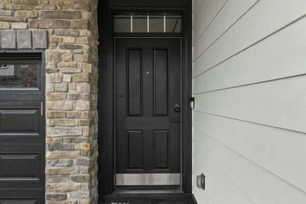 A black front door with a small window above it is set between a stone wall and white horizontal siding, next to a black garage door.
