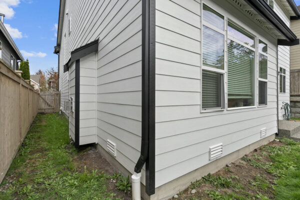 A narrow side yard with a grass strip runs alongside a modern light gray house with white trim and multiple vents. A wooden fence and gate border the yard; a concrete pathway is visible on the right.