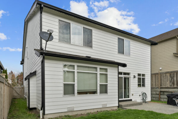 A two-story modern house with white siding, black trim, and several windows, featuring a small concrete patio in the fenced backyard on a clear, sunny day.