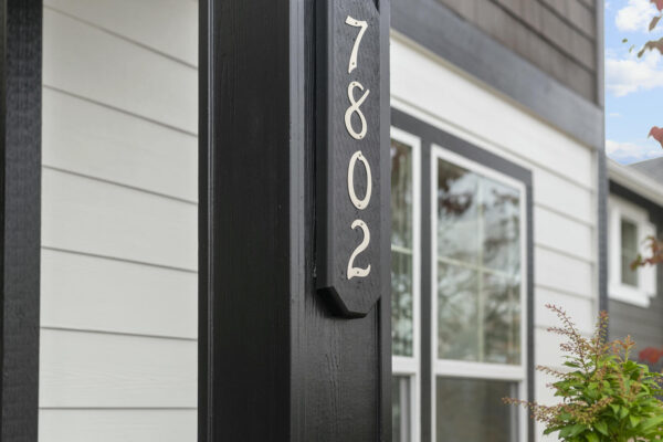 A black pillar with the house number 7802 in white, in front of a modern house with white and gray siding, large windows, and some greenery nearby.