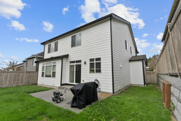 A modern two-story white house with large windows, a small patio featuring a covered grill and a power washer, and a fenced grassy backyard under a blue sky with scattered clouds.