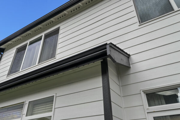 Close-up of the exterior of a two-story house with white horizontal siding, black trim, and several windows, set against a blue sky with a few clouds.