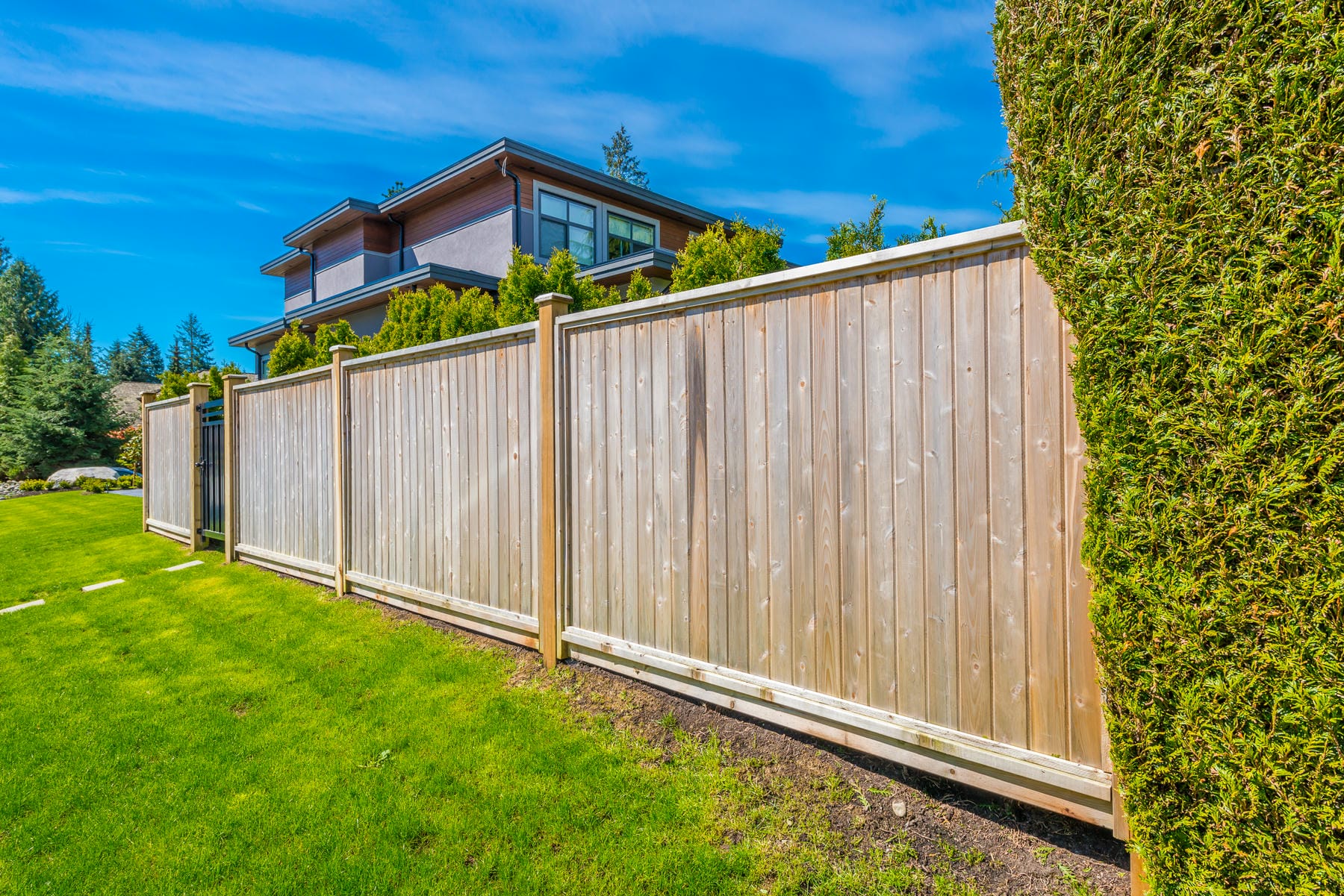 A tall wooden privacy fence stands along a well-maintained green lawn, with a modern two-story house and trimmed hedges visible in the background under a bright blue sky.