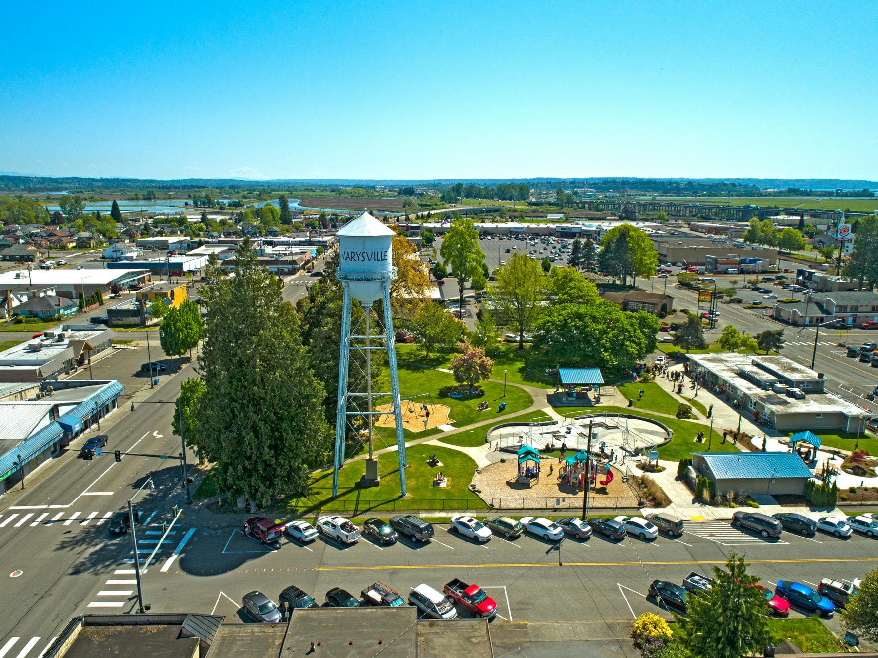 Aerial view of a park with a playground, green trees, and a water tower labeled Mossville, surrounded by parking lots, cars, and commercial buildings under a clear blue sky.