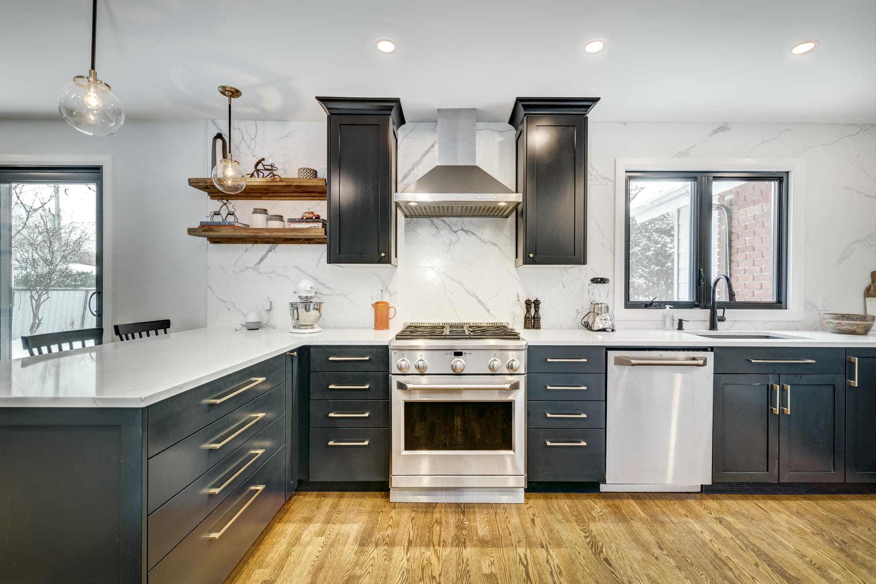 Modern kitchen with black cabinets, stainless steel stove, dishwasher, range hood, white marble backsplash, wood floating shelves, and a farmhouse sink under a window. The floor is wood, and countertops are white.
