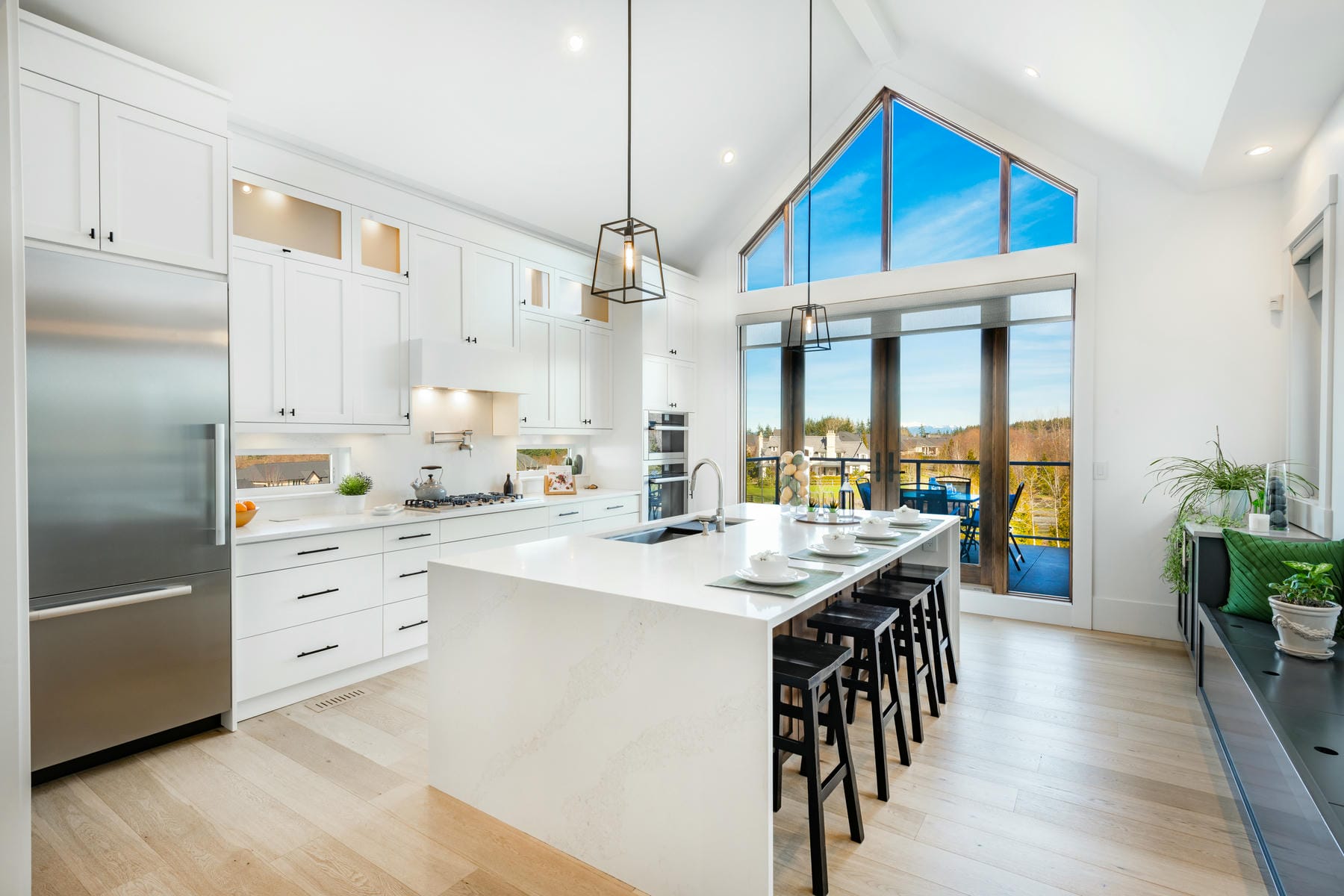 Modern, bright kitchen with white cabinets, a large marble island with bar stools, stainless steel appliances, and floor-to-ceiling windows overlooking a deck and scenic outdoor view.