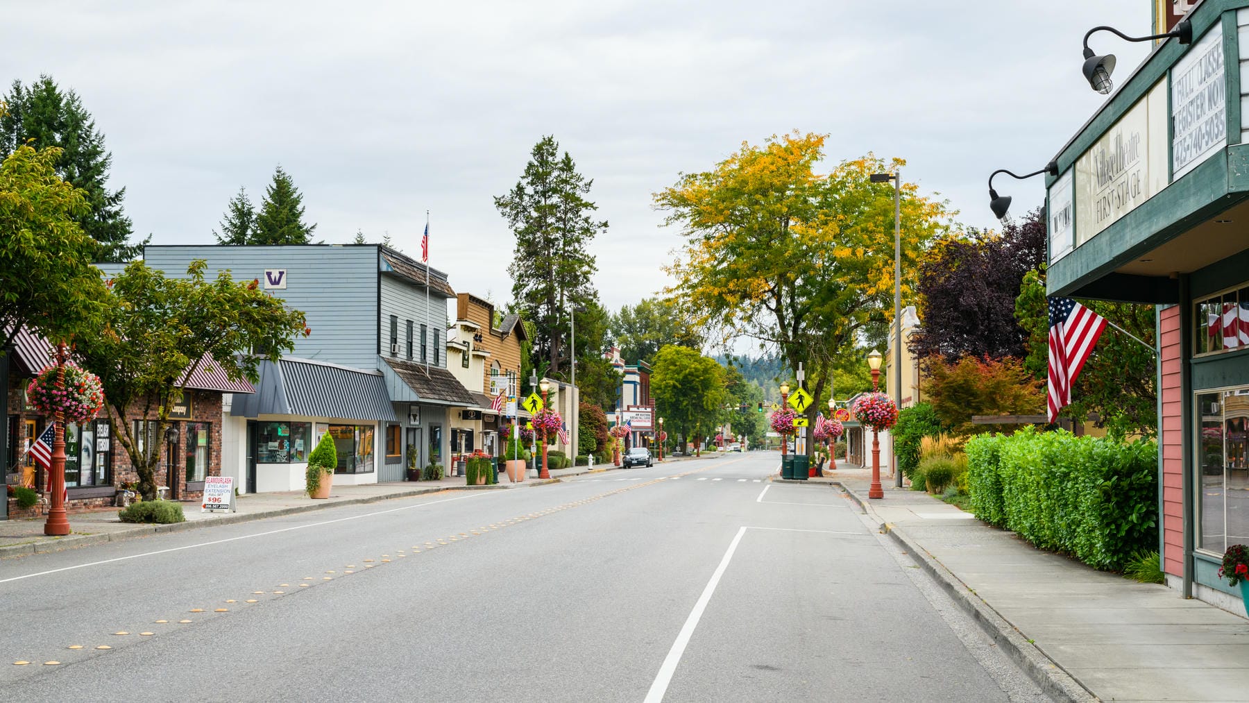 A quiet small-town street lined with shops, hanging flower baskets, trees, and American flags on a cloudy day. There are no people or cars visible, and the scene appears calm and well-kept.
