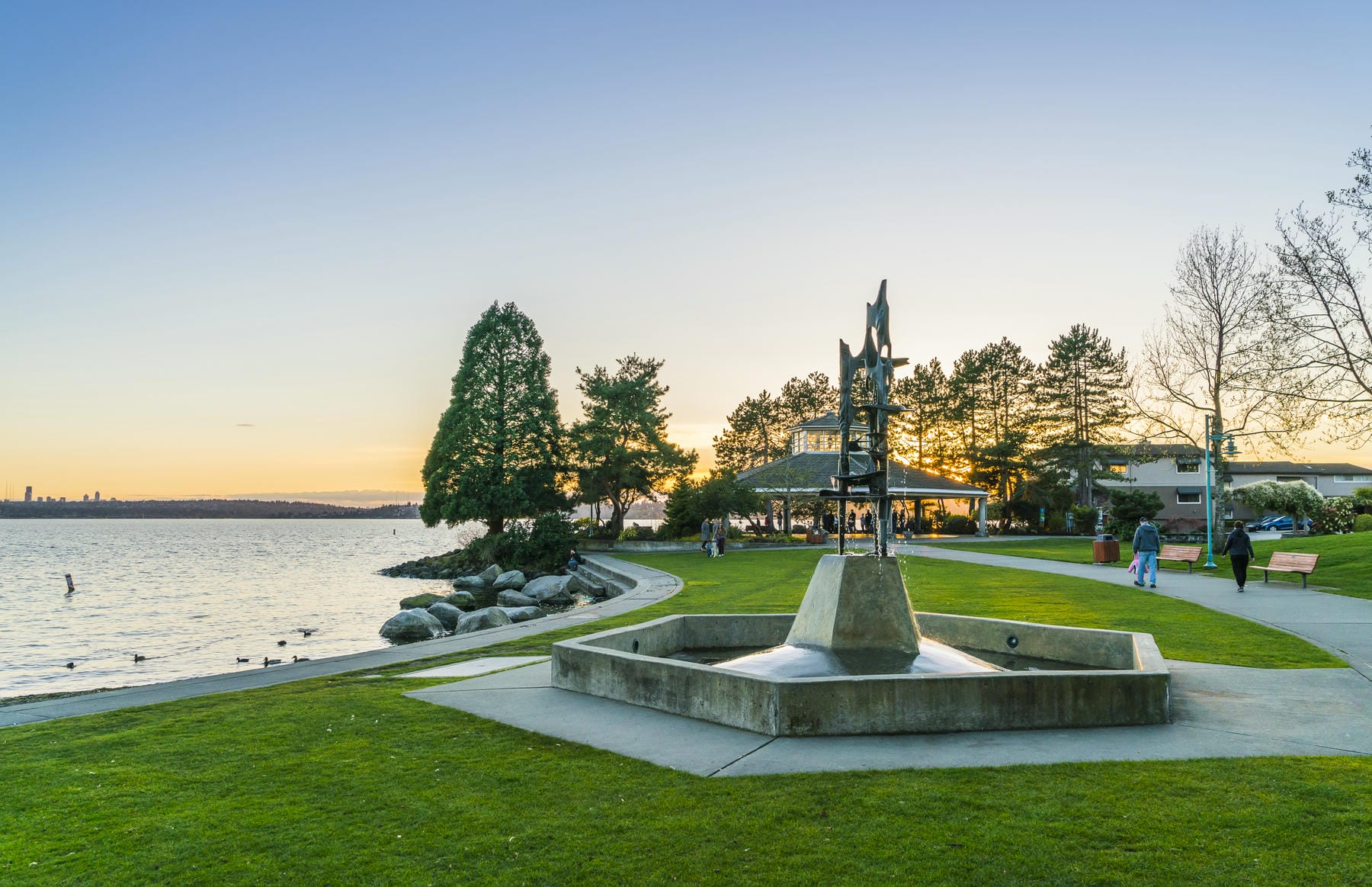 A lakeside park at sunset with a modern sculpture on a concrete base in the foreground, a gazebo surrounded by trees, people walking, and water with distant city skyline in the background.