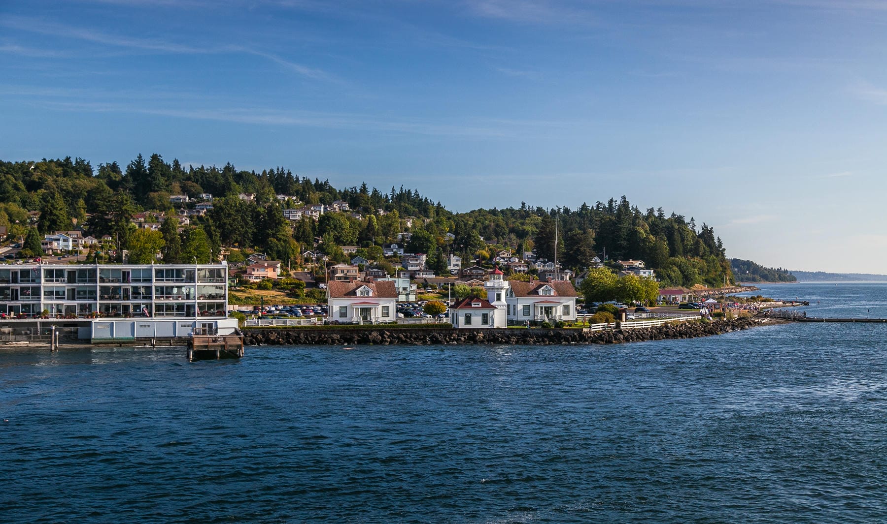 A coastal scene shows a small peninsula with white buildings, including a lighthouse, surrounded by blue water. Modern apartments and houses sit among green trees on a hillside under a clear sky.