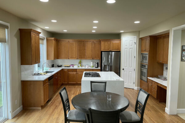 A spacious modern kitchen features wooden cabinets, stainless steel appliances, and a white island with a built-in cooktop. A round dining table with four chairs sits on a hardwood floor. The room is well-lit with recessed ceiling lights.