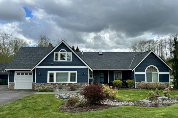 A dark blue house with white trim and a stone facade sits on a green lawn with shrubs and rocks, under a cloudy sky with trees in the background.