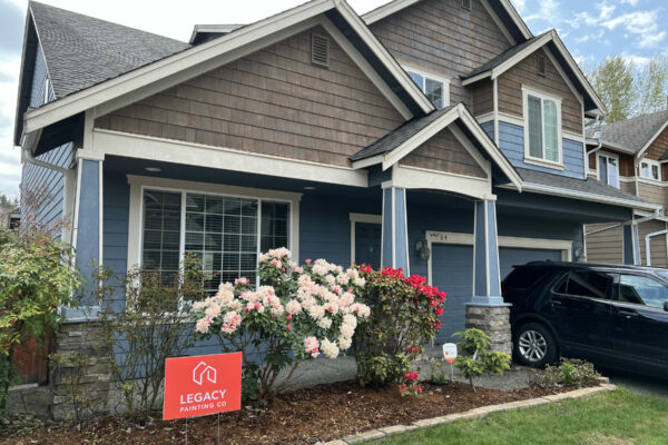 A two-story house with blue siding and brown shingles, white trim, and a front garden with blooming flowers. A black SUV is parked in the driveway, and a red Legacy Painting Co sign is placed in the yard.