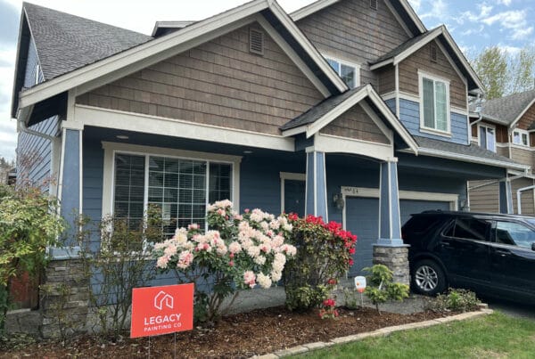 A two-story house with blue siding and brown shingles, white trim, and a front garden with blooming flowers. A black SUV is parked in the driveway, and a red Legacy Painting Co sign is placed in the yard.