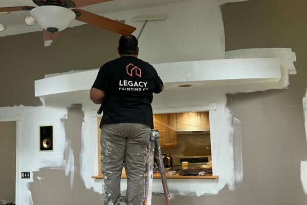A painter stands on a ladder, using a roller to paint a brown wall white. He wears a black Legacy Painting shirt and paint-stained pants. Paint supplies and drop cloths are on the floor; the room features a ceiling fan and track lights.