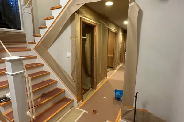A staircase and hallway in a house with floors, stairs, and walls covered in brown protective paper and tape, likely for painting or renovations. A blue container and various tools are on the floor.