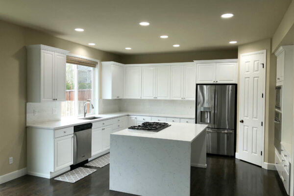 Modern kitchen with white cabinets, stainless steel appliances, a large island with a cooktop, and dark wood floors. Recessed ceiling lights illuminate the space, and a window provides natural light near the sink.