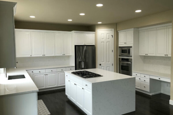 Modern kitchen with white cabinets, a large island featuring a gas cooktop, built-in stainless steel appliances, dark wood flooring, and recessed ceiling lights. The space is clean and well-lit.