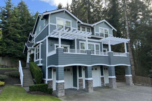 Two-story, blue-gray house with white trim and pergolas over balconies, three garage doors, surrounded by trees and landscaping, with a paved driveway and green lawn.