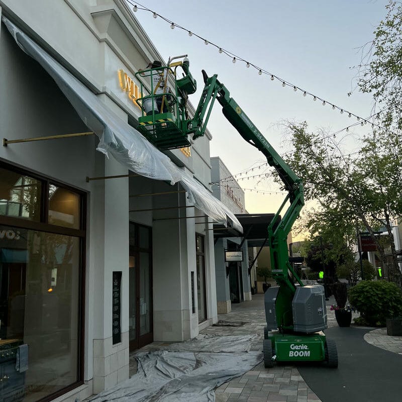 A worker stands on a green Genie boom lift, working near the roof of a building covered in plastic sheeting, with string lights overhead and trees lining the sidewalk below.