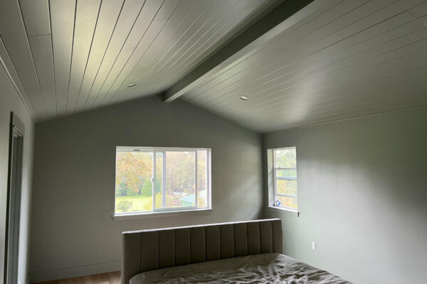 A minimally furnished bedroom with light gray walls and ceiling, a large window with a view of trees, a small side window, and an upholstered bed with rumpled bedding. Natural light fills the room.