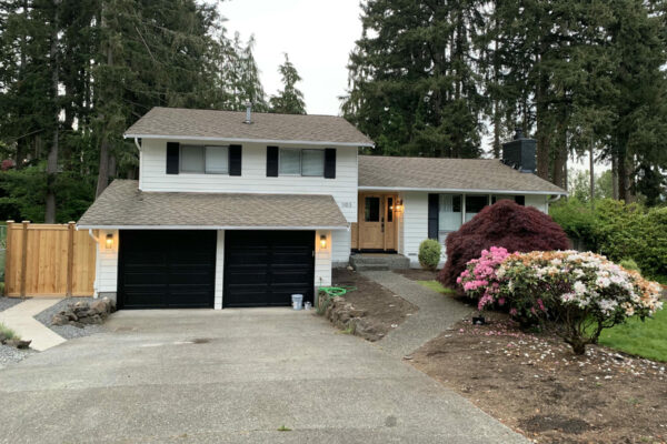 A two-story suburban house with white siding, black shutters, and a two-car garage with black doors. There is a paved driveway, a small front porch, and landscaped bushes and flowers along the walkway.