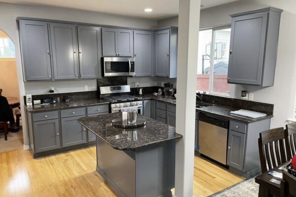 Modern kitchen with gray cabinets, stainless steel appliances, granite countertops, and a small island. Light-colored wood floor and a dining area with a dark wooden table and chairs are visible. The space is well-lit with overhead lights.