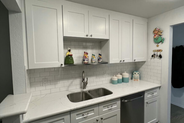 A modern kitchen with white cabinets, a double sink, stainless steel dishwasher, white tile backsplash, colorful decor above the cabinets, and a patterned rug on the gray wood floor.