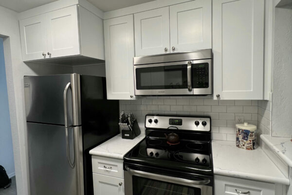 Modern kitchen with white cabinets, stainless steel refrigerator, oven, and microwave. Countertops are white with a white subway tile backsplash. There is a knife block and a decorative container on the counter.