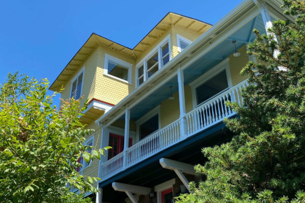 A bright yellow two-story house with a white porch and blue ceiling is surrounded by lush green plants and trees under a clear blue sky.