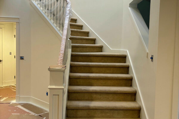 A staircase with beige carpet is flanked by white railings. The walls are painted a light color, and the area appears to be under renovation, with protective paper covering parts of the floor and railing.