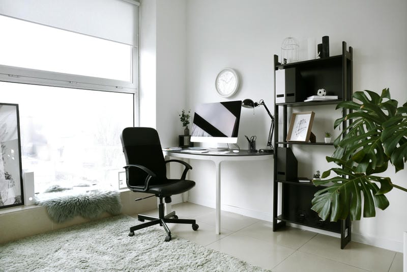 Minimalist home office with a black chair, desk, computer, bookshelf, wall clock, plants, and large window with natural light. Green rug and a cozy seat are by the window. Decor is modern with a clean, white aesthetic.