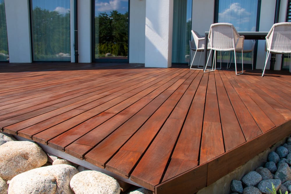 A close-up view of a wooden patio deck with smooth, reddish-brown planks next to a modern house. White chairs and a table are on the deck, and there are large round stones along the edge.