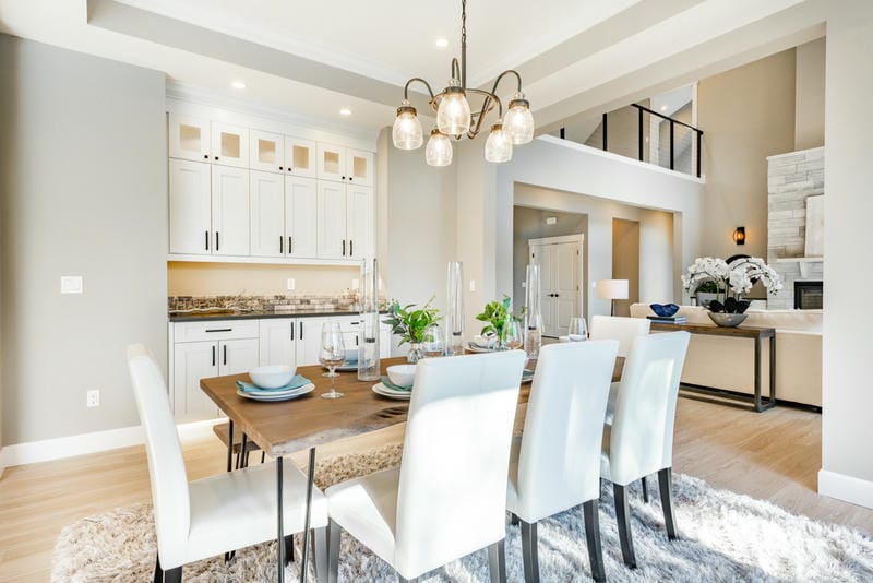 A modern dining room with a wooden table set for six, white chairs, light wood flooring, a soft rug, pendant lighting, and an open layout revealing a kitchen and living area with neutral tones.