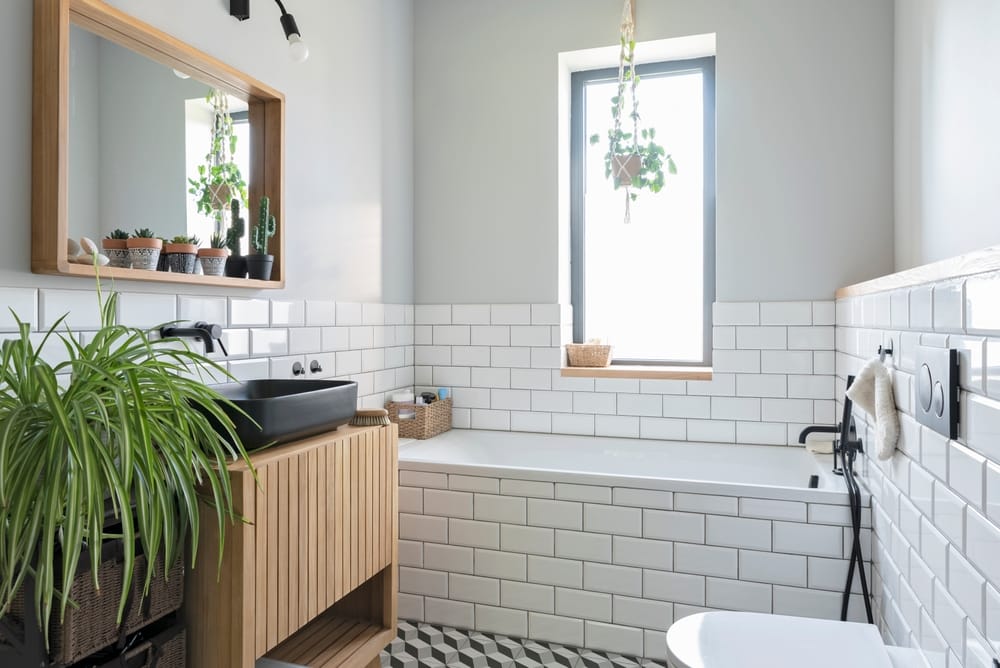 Modern bathroom with white subway tiles, a wooden vanity with a black sink, a large mirror, potted plants, a bathtub under a window, and patterned floor tiles. Hanging plants add a fresh touch.