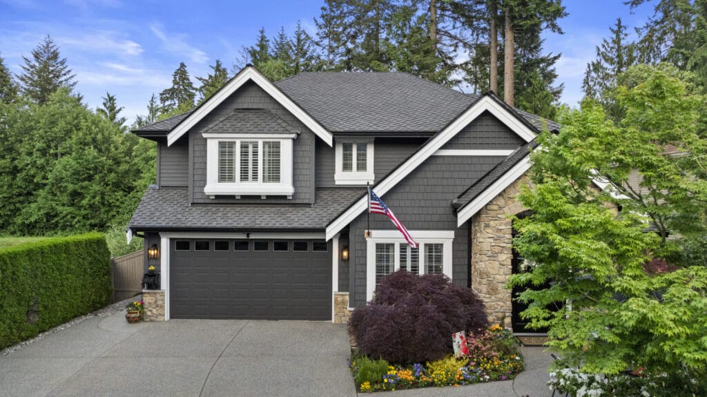 A two-story modern house with dark gray siding, stone accents, and a double garage. An American flag hangs near the front door, surrounded by landscaped flowers and trees, with tall evergreens in the background.