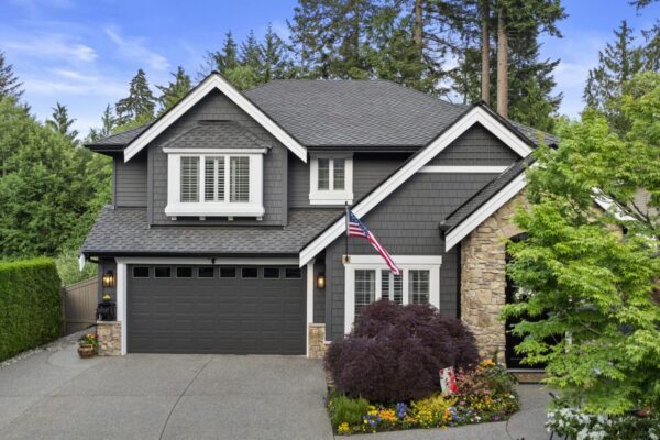 A two-story modern house with dark gray siding, stone accents, and a double garage. An American flag hangs near the front door, surrounded by landscaped flowers and trees, with tall evergreens in the background.