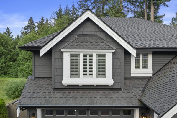Close-up of a house exterior featuring dark gray siding, white window trim, shuttered windows, and a gabled roof with asphalt shingles, set against a backdrop of green trees and a blue sky.
