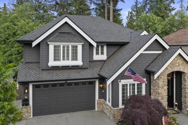 Two-story gray house with white trim, stone accents, and an American flag by the front entrance. Surrounded by trees and greenery, the house has a double garage and manicured landscaping.