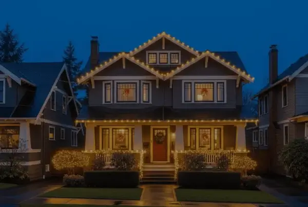 A two-story house decorated with warm white string lights along the roofline and bushes, with lit wreaths in the windows, sits between two similar homes at dusk. The sidewalk and street in front appear wet.