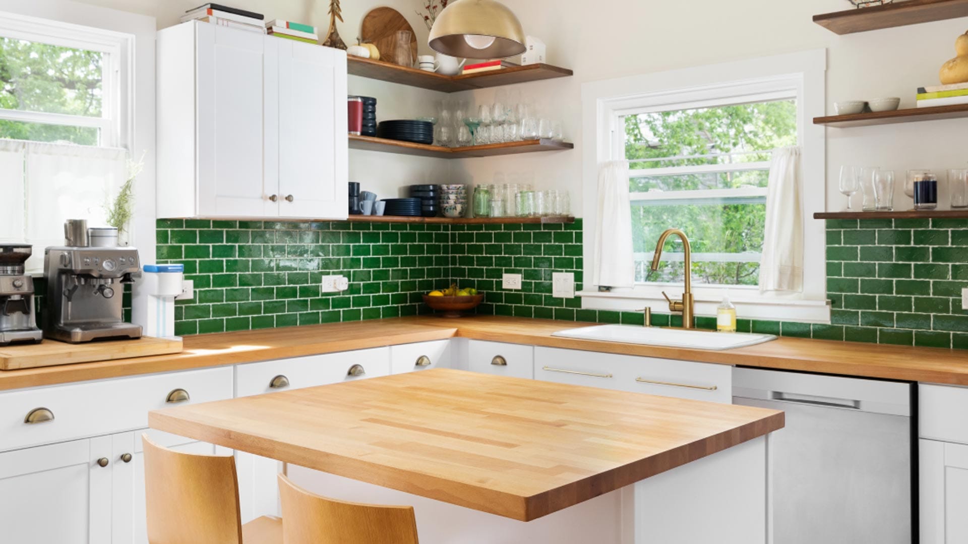 Modern kitchen with white cabinets, wooden countertops, green subway tile backsplash, open shelving with dishes, a gold faucet, and a coffee machine by the window. Two wooden chairs are at the center island.
