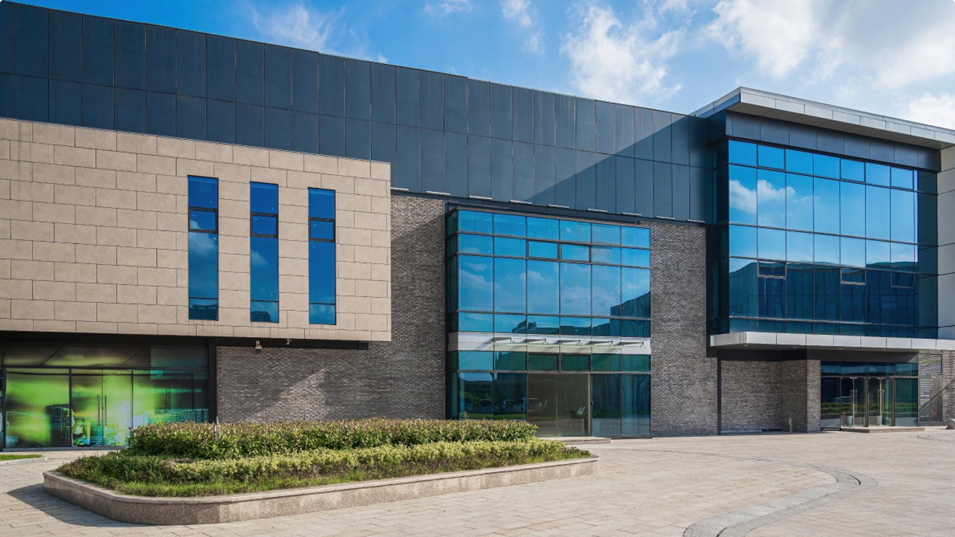 A modern commercial building with large blue-tinted glass windows, beige brick and dark gray panels, set against a partly cloudy sky with landscaped greenery in front.