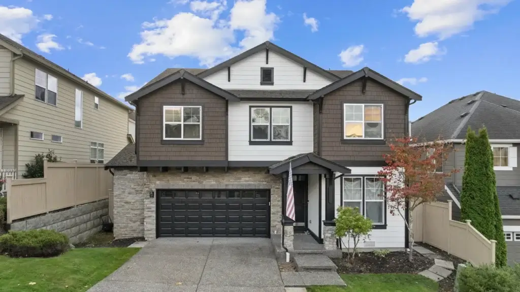 Two-story modern suburban house with brown and white siding, stone accents, black garage door, small front porch, American flag, and landscaped yard, set between two other houses under a partly cloudy sky.