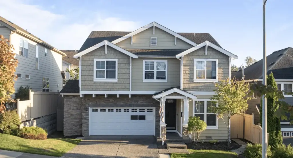 Two-story beige suburban house with white trim, stone accents, and a two-car garage. There are trees and shrubs in the yard, a paved driveway, and neighboring houses on both sides under a clear blue sky.