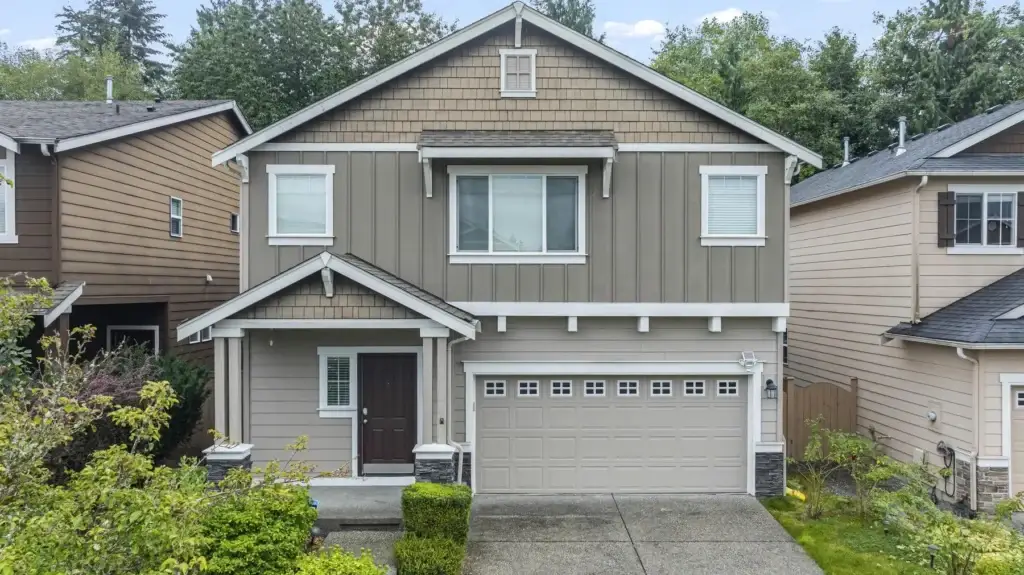Two-story suburban house with taupe siding, white trim, a double garage, and a covered front porch. The house is surrounded by greenery and neighboring homes, with a concrete driveway and walkway.